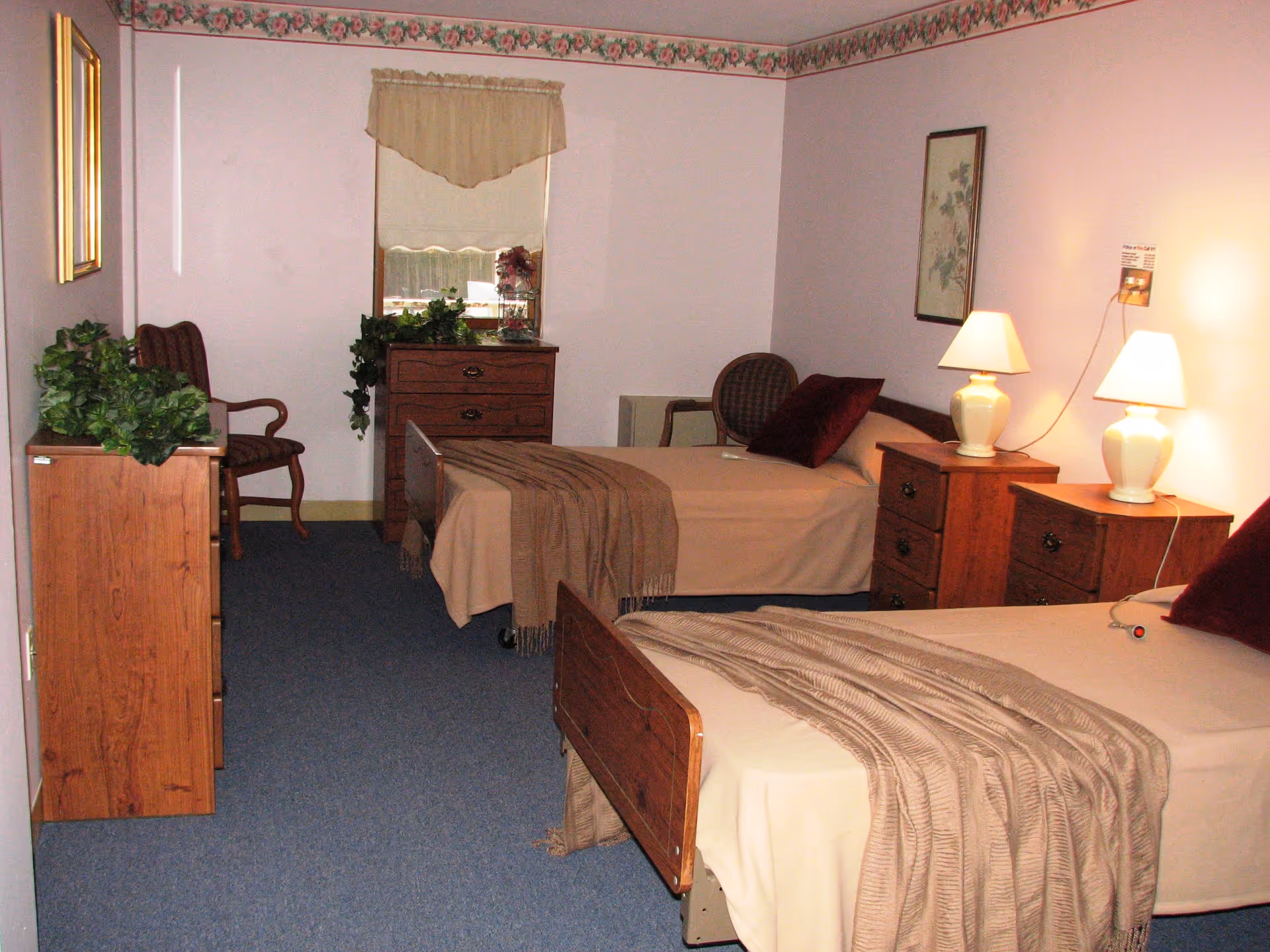 A bedroom with two single beds covered in beige bedspreads and brown throws. Each bed has a burgundy pillow. There are two wooden nightstands with white lamps between the beds. A wooden dresser with plants and a chair are against the left wall. A window with a beige valance is on the far wall, and a framed picture hangs on the right wall. The room has a blue carpet and light pink walls with floral border wallpaper near the ceiling.