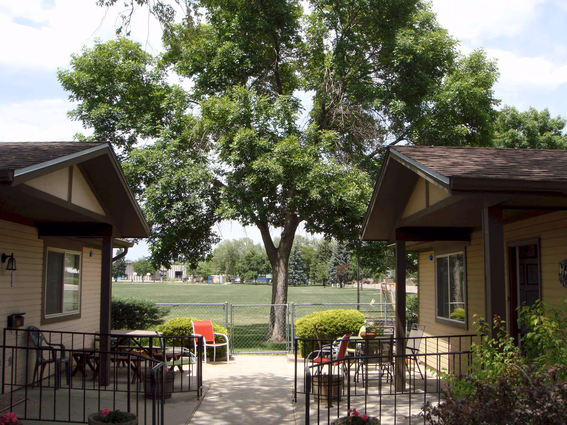 Courtyard between two single-story senior housing units with patio chairs and tables, a large tree and a grassy field beyond a chain-link fence.