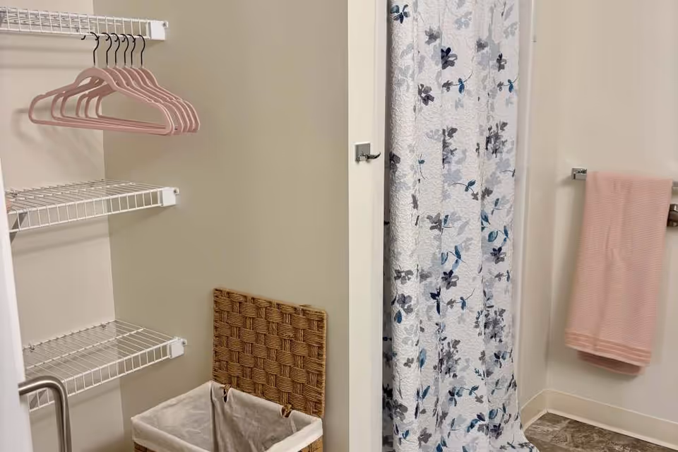 A bathroom corner with a floral shower curtain, a pink towel hanging on a towel rack, white wire shelves with pink hangers, and a woven laundry basket with a fabric liner.