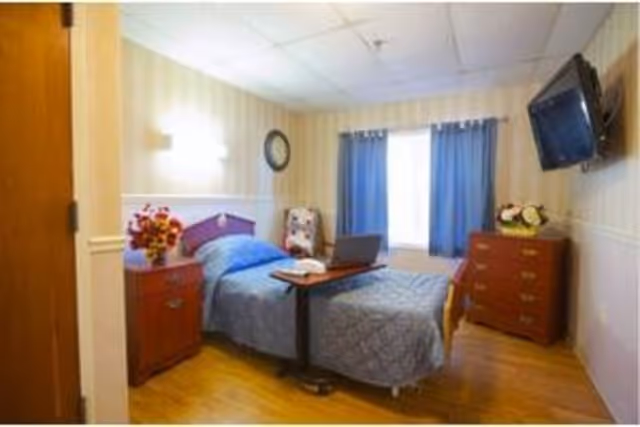 Well-lit bedroom with a single bed, bedside tables with flowers, a wall-mounted TV, and blue curtains.
