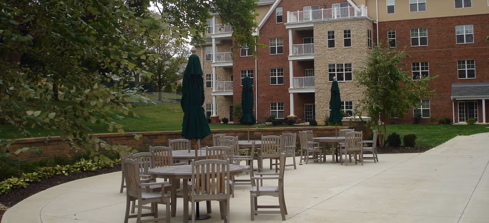 Outdoor patio area with multiple round wooden tables and chairs, each table having a closed green umbrella. The patio is adjacent to a multi-story brick building with balconies and windows. There is greenery including trees and shrubs surrounding the patio.