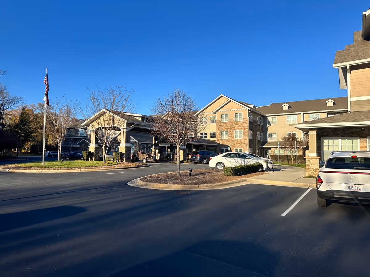 Front exterior of a multi-story senior living facility with an entrance canopy, parked cars, and an American flag under a clear blue sky.