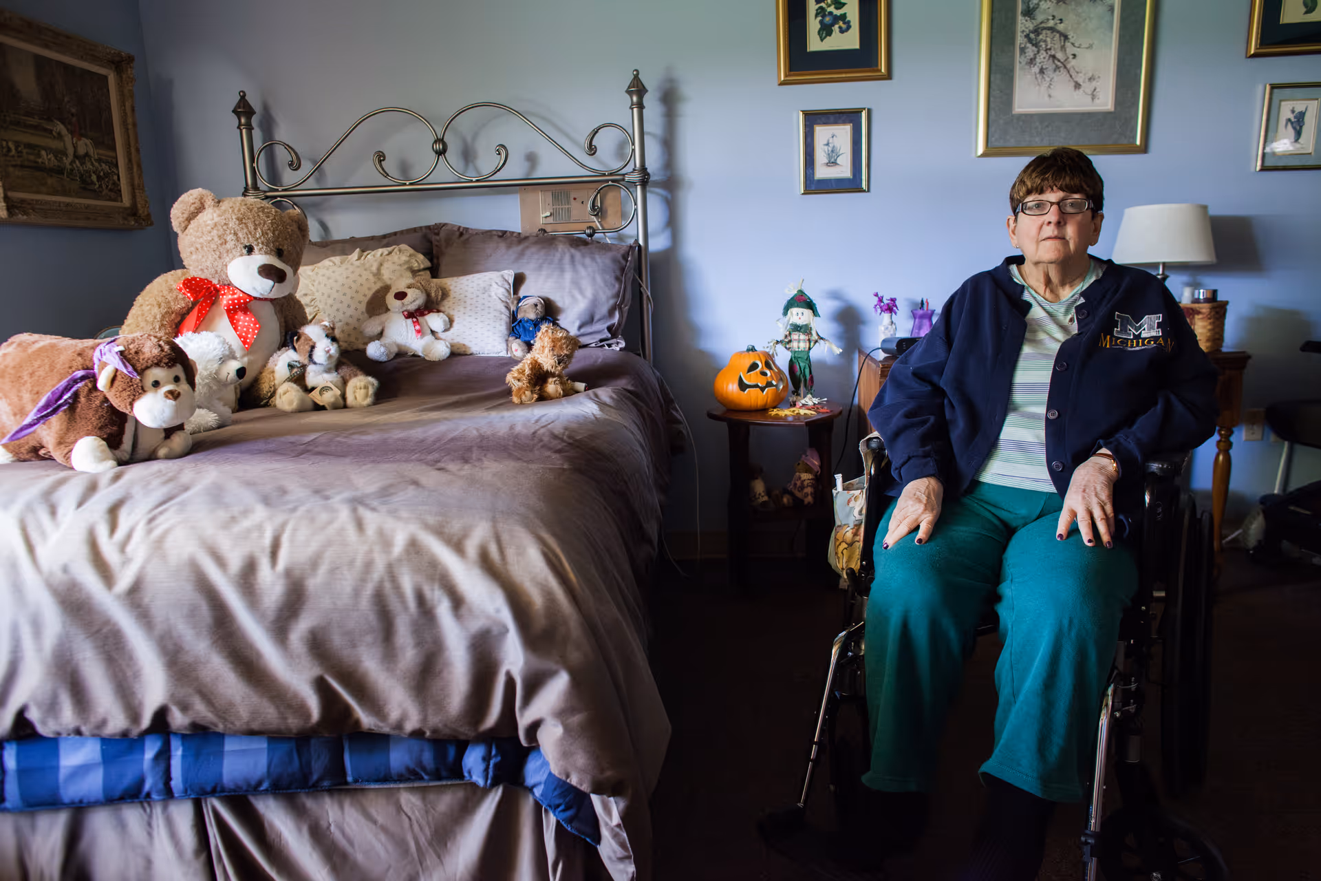 An older woman in a wheelchair sits next to a bed covered with stuffed animals and pillows in a bedroom.