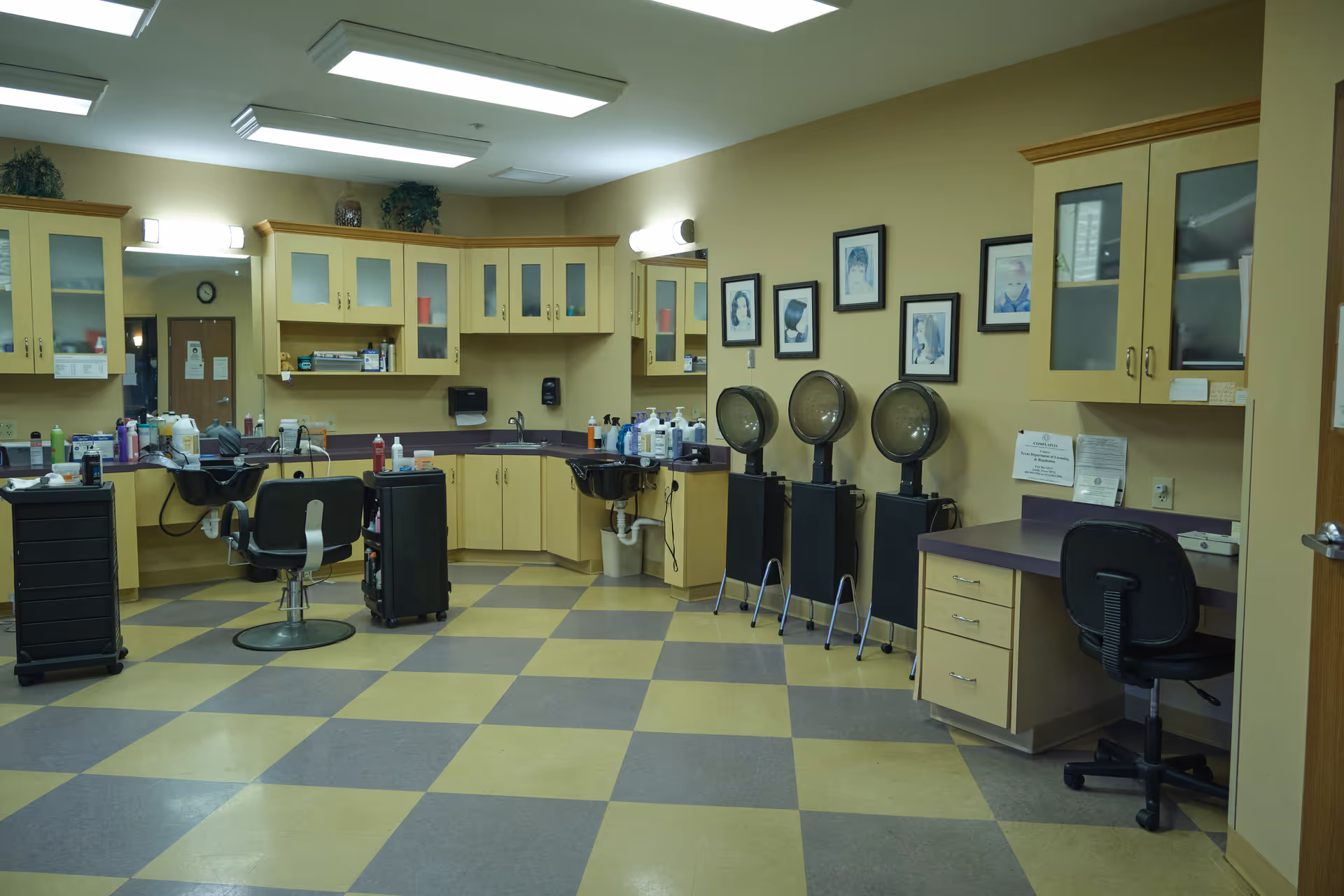 Interior of a salon area in a senior living facility with salon chairs, hair washing sinks, hair dryers, cabinets, and framed pictures on the wall. The floor has a checkered pattern with yellow and gray tiles.