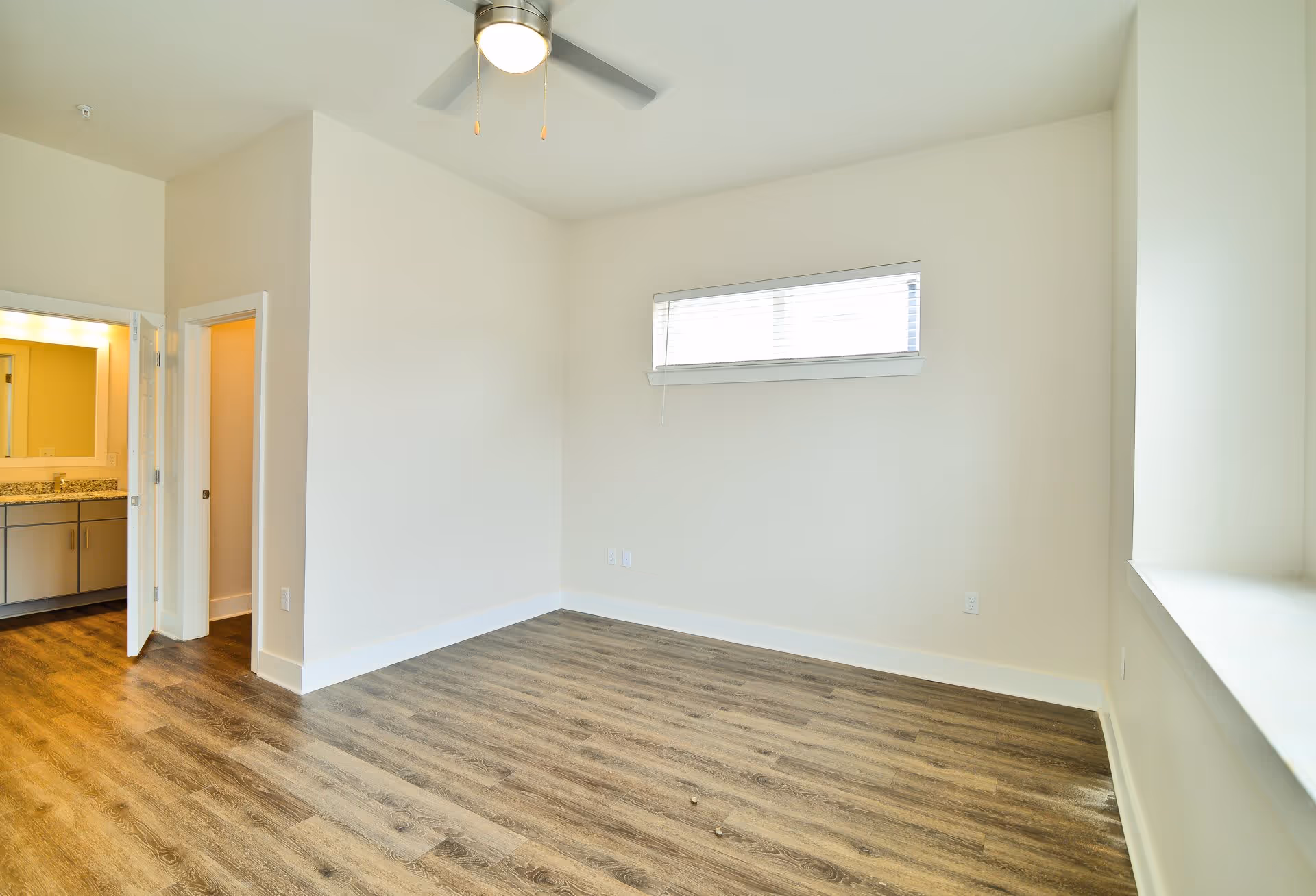 Empty room with wood flooring, white walls, a ceiling fan with light, a narrow horizontal window with blinds, and an open door leading to a bathroom with a sink and mirror.