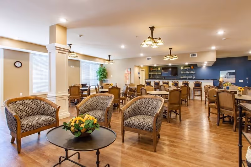A spacious and well-lit common area with wooden flooring, featuring multiple tables and chairs arranged for dining or socializing. In the foreground, there are three patterned armchairs around a round coffee table with a flower arrangement. The back wall has a bar area with stools, shelves with bottles, and a mounted flat-screen TV. Large windows with blinds allow natural light to fill the room, and ceiling lights provide additional illumination.