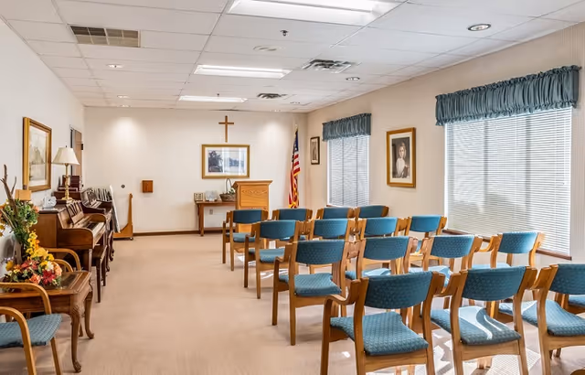 Sunlit chapel-style meeting room with rows of teal-upholstered chairs facing a wooden podium and a cross on the wall.