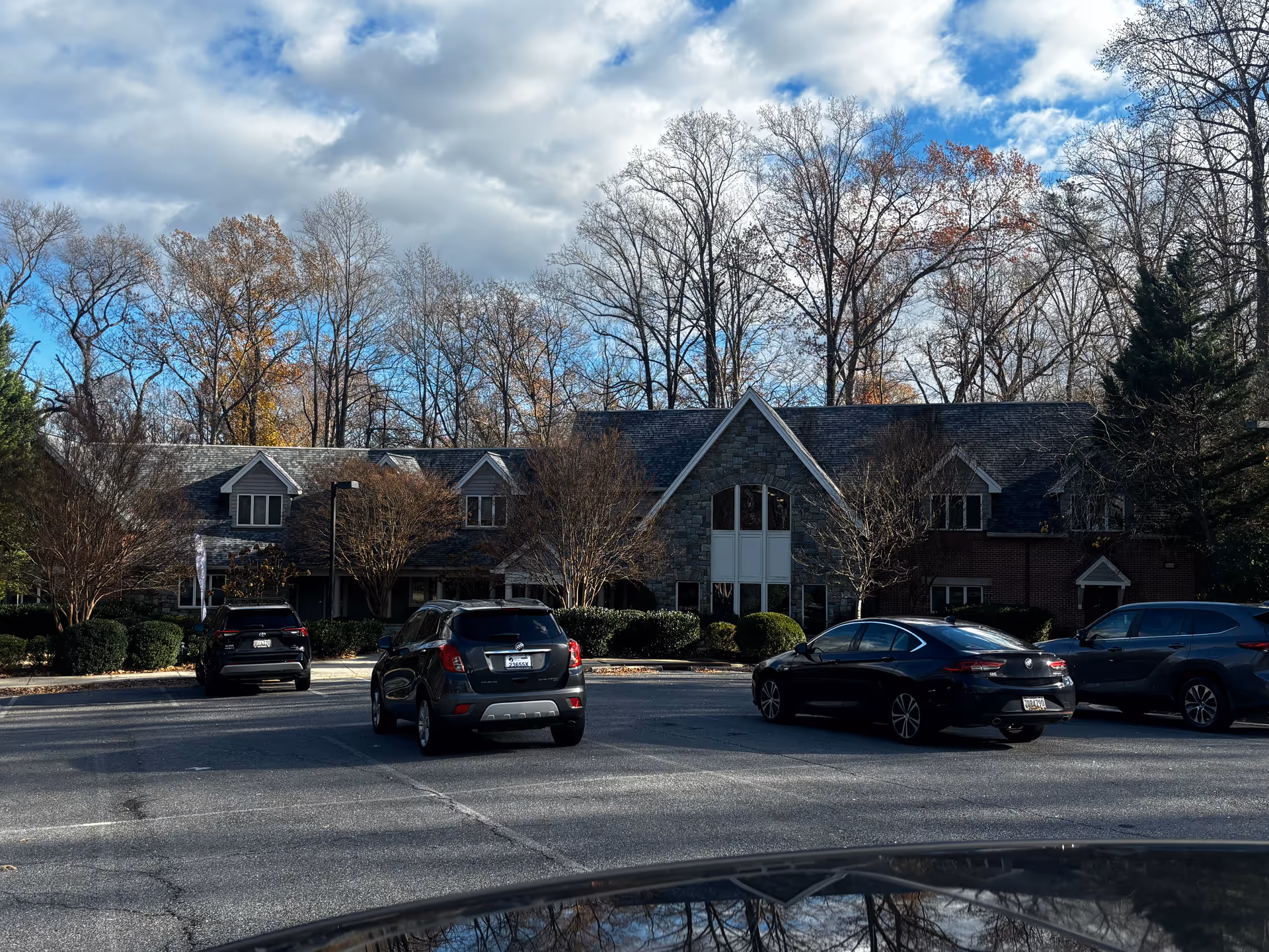 Exterior view of a senior living facility named Byron House, showing a large building with stone and brick facade, multiple windows, and a sloped roof. Several cars are parked in the parking lot in front of the building, with leafless trees and a partly cloudy sky in the background.