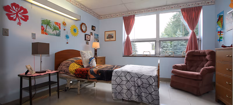 A bright senior living bedroom with a single adjustable bed, bedside tables and lamp, a pink upholstered armchair, large window with pink curtains, and colorful wall decorations.