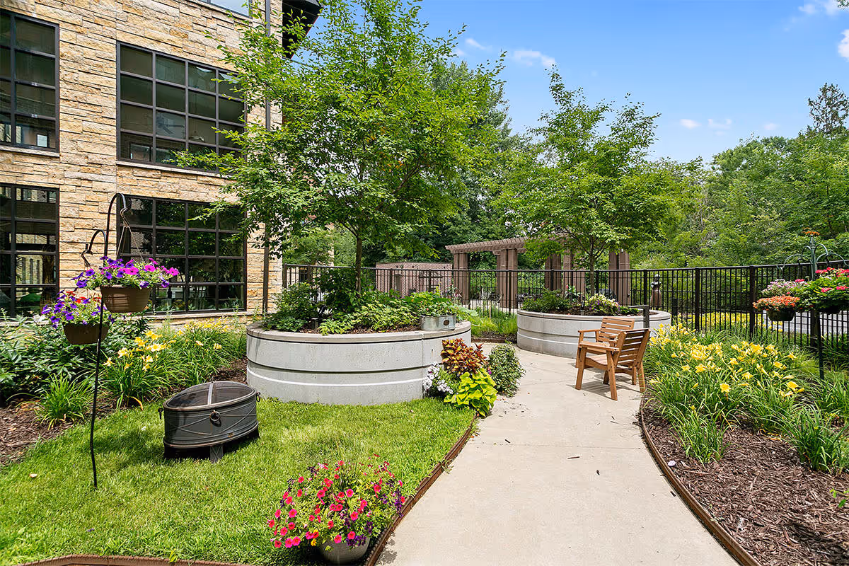 Outdoor garden area at a senior living facility with a paved walkway, two wooden chairs, raised circular planters with trees and flowers, hanging flower baskets, and a stone building with large windows in the background.