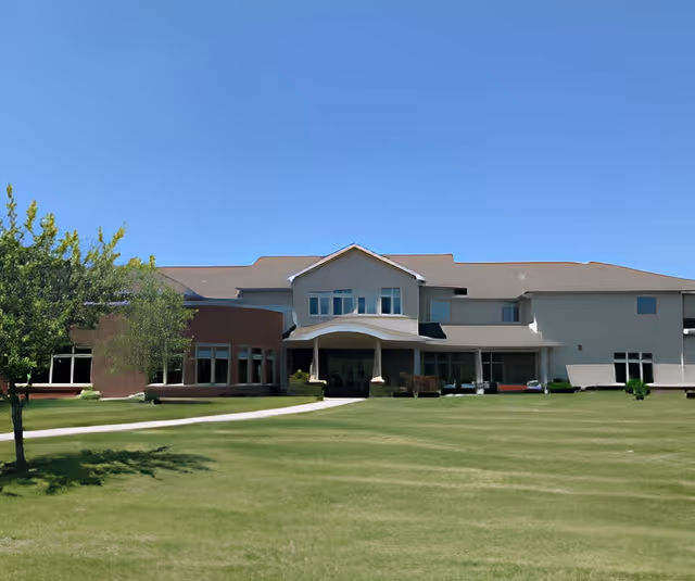 Front exterior of a two-story senior living building with a covered main entrance, expansive lawn, and clear blue sky.