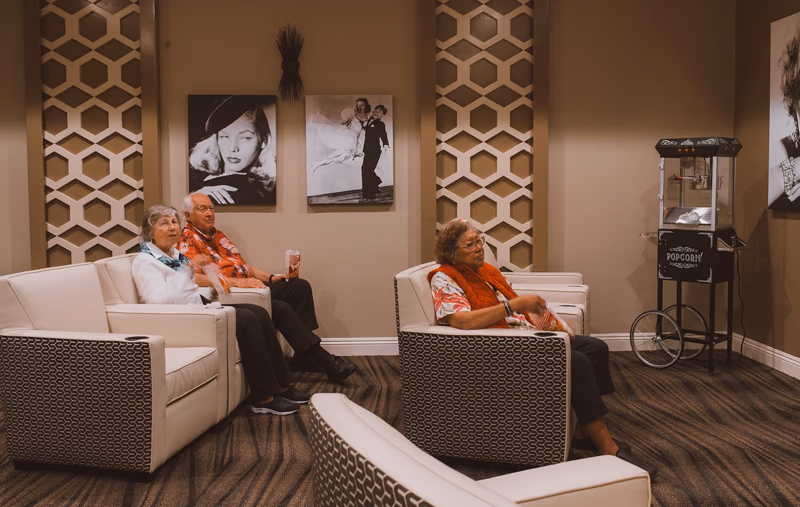 Three elderly people sitting in a cozy lounge area with patterned armchairs and a popcorn machine. The walls are decorated with black and white vintage-style portraits and geometric wall panels.