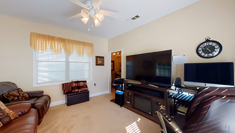 A cozy living room with beige walls and carpet, featuring a brown leather sofa with patterned pillows on the left. A large flat-screen TV sits on a dark wooden entertainment center with a fireplace insert. A ceiling fan with lights is mounted on the ceiling. There is a window with a yellow valance letting in natural light. A clock hangs on the wall above a computer monitor on the right side of the room.