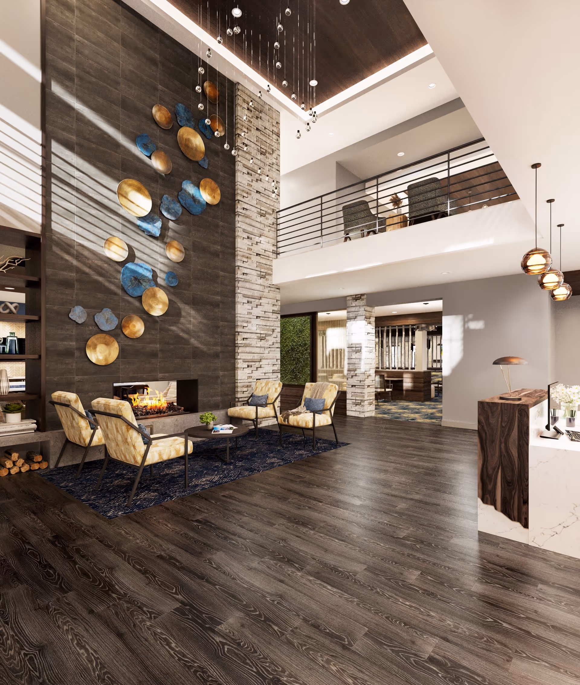 A modern and spacious living room area with dark wood flooring, a tall stone fireplace with a fire burning, and decorative wall art featuring gold and blue circular elements. Four cushioned chairs with yellow patterned upholstery are arranged around a small round coffee table on a dark blue rug. The room has a high ceiling with hanging pendant lights and a mezzanine level with additional seating visible. A reception desk with a lamp and flowers is on the right side of the image.