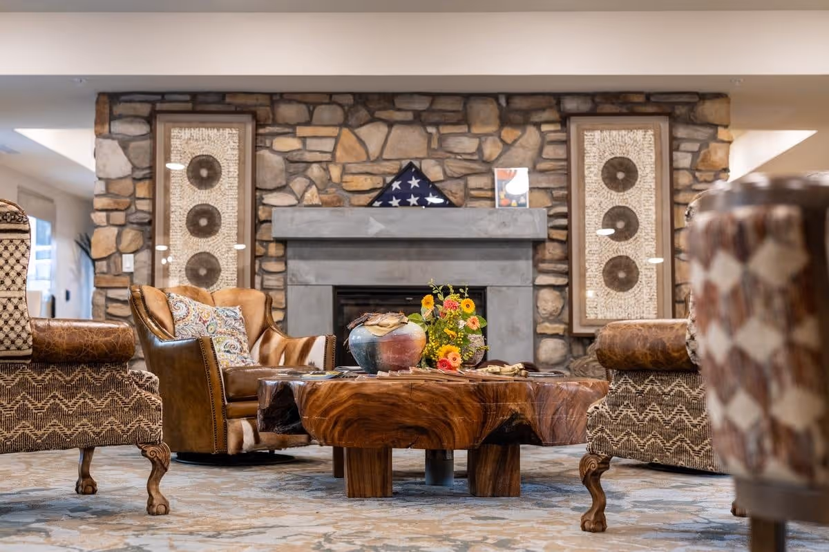 Cozy senior living common area with a stone fireplace, a wooden coffee table with decorative items and flowers, surrounded by comfortable upholstered armchairs and leather chairs, with framed artwork on the stone wall.