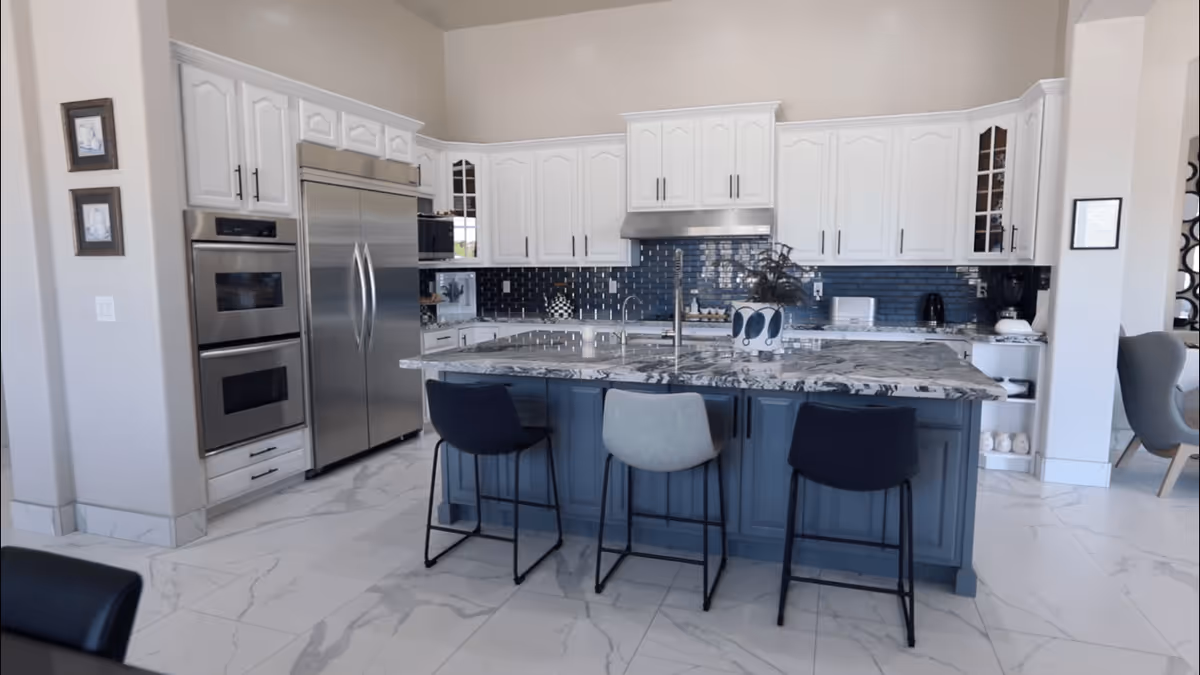 Spacious modern kitchen with a large marble island, three bar stools, stainless steel refrigerator and ovens, and white cabinetry.