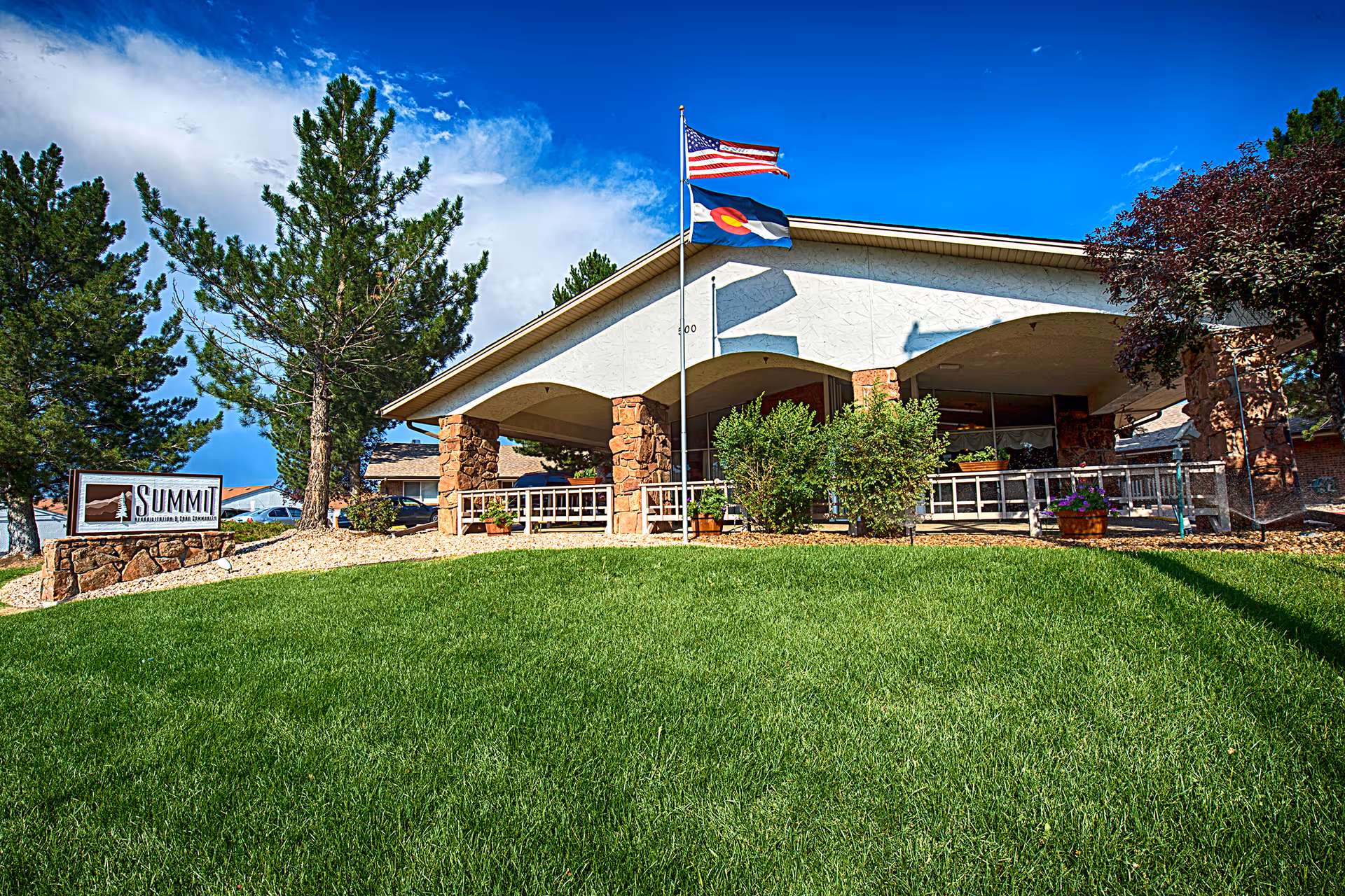 Front exterior of a single-story care facility with an American and Colorado flag, a green lawn, and a 'Summit' sign.