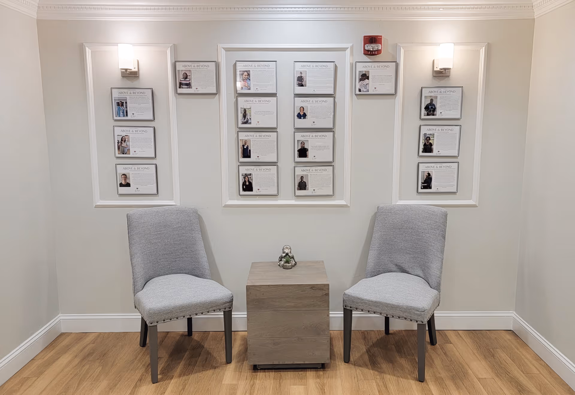 A small seating area with two gray upholstered chairs on either side of a wooden side table with a small decorative figurine. Behind the chairs, there are three wall panels each displaying framed photos and text titled 'Above & Beyond'. The walls are light-colored, and there are two wall-mounted lights above the panels.