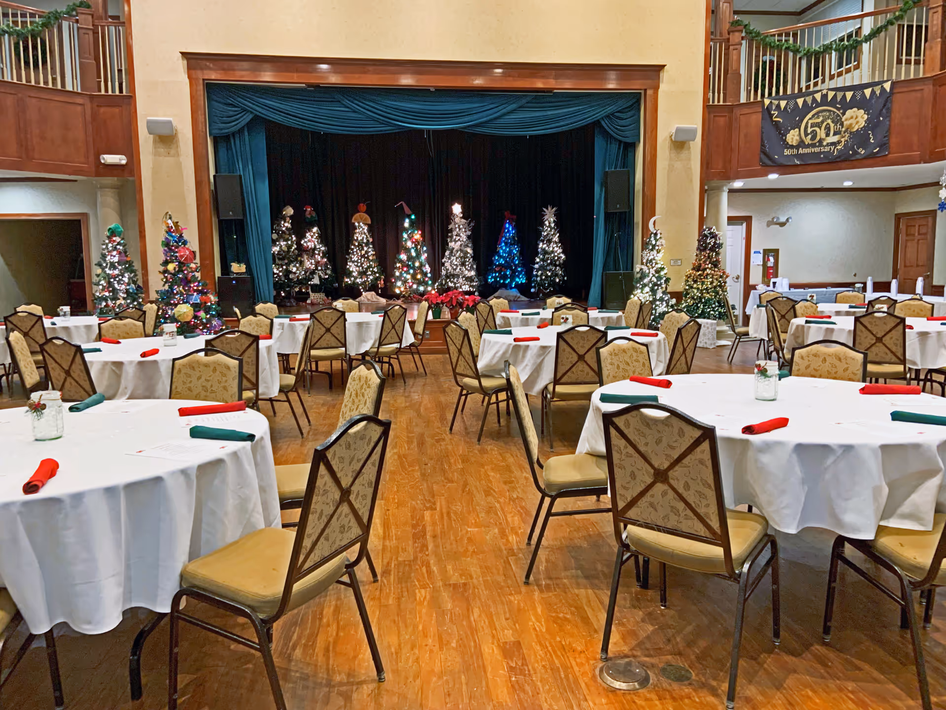 A festive dining or event room with round tables covered in white tablecloths and chairs arranged around them. Each table has red and green folded napkins and small decorative centerpieces. In the background, there is a stage with multiple decorated and lit Christmas trees. The room has wooden flooring and a balcony area above with a banner celebrating a 50th anniversary.