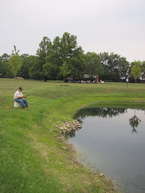 A man sitting on a bucket fishing by the edge of a small pond surrounded by green grass and trees in the background under a cloudy sky.