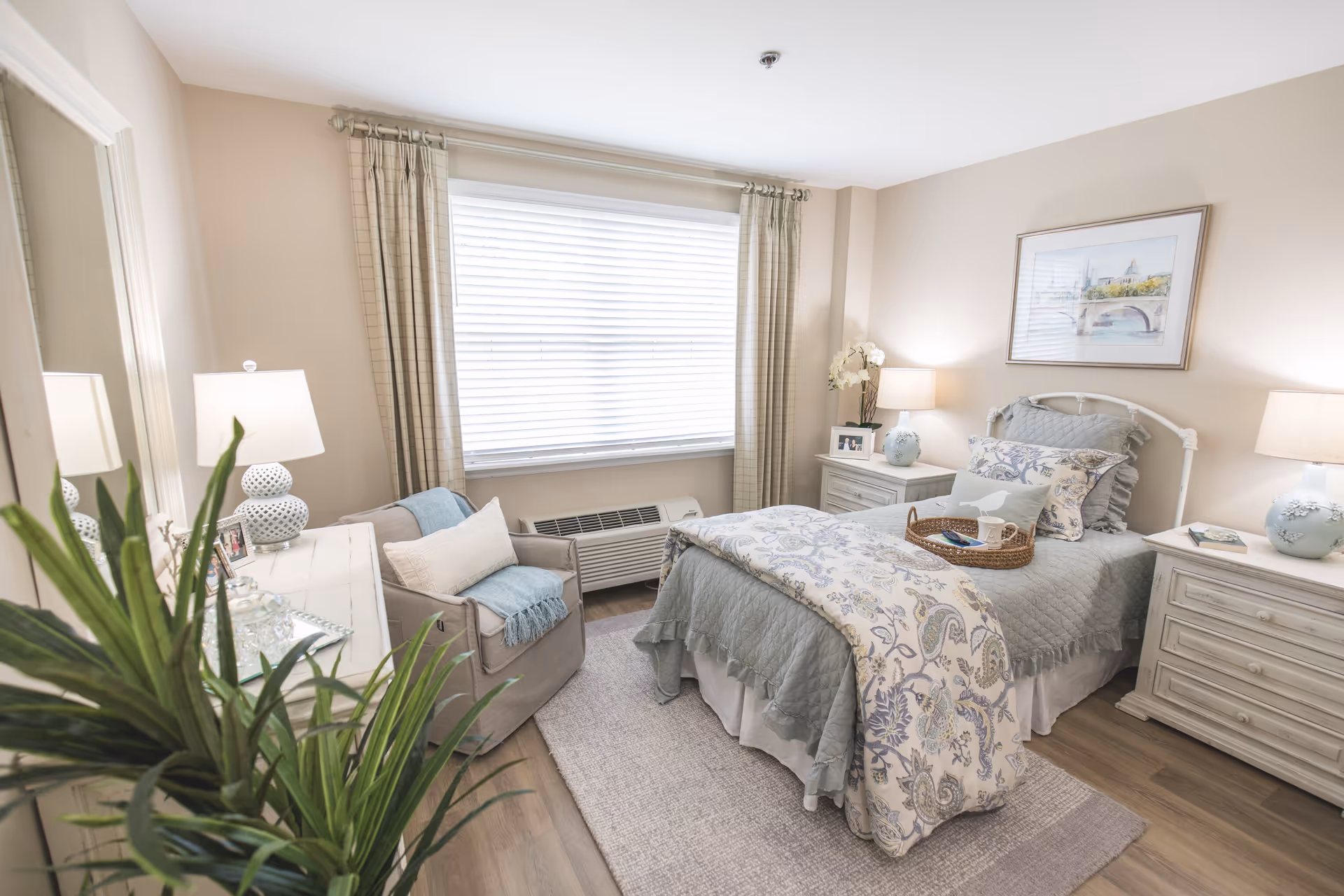 A cozy bedroom with a single bed covered in a light blue quilt and a floral patterned blanket. There are two matching nightstands on either side of the bed, each with a lamp and decorative items. A comfortable armchair with a pillow and throw blanket is positioned near a large window with closed blinds and beige curtains. A large mirror and a green plant are visible in the foreground, and the room has light-colored walls and wooden flooring.