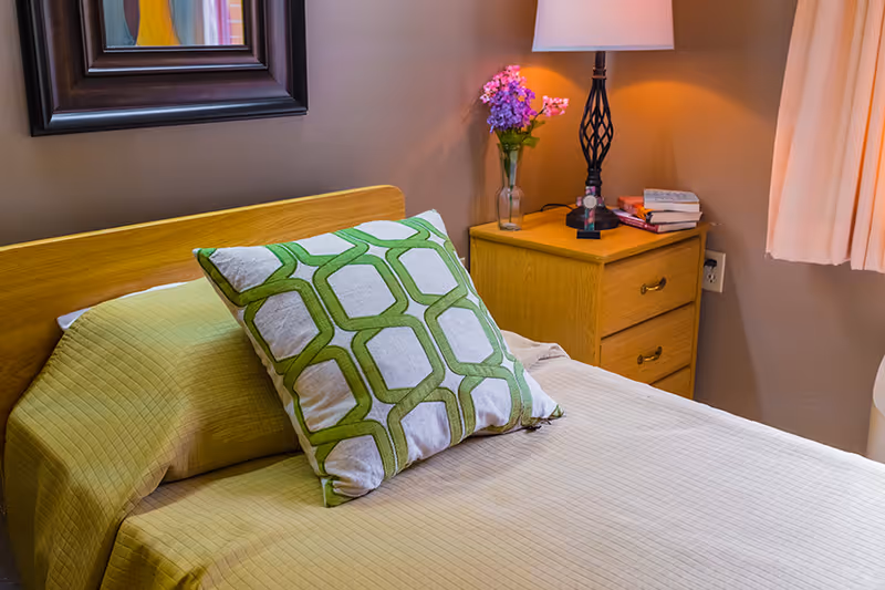 Neatly made single bed with a green patterned pillow, a wooden headboard, and a nightstand with a lamp and flowers.