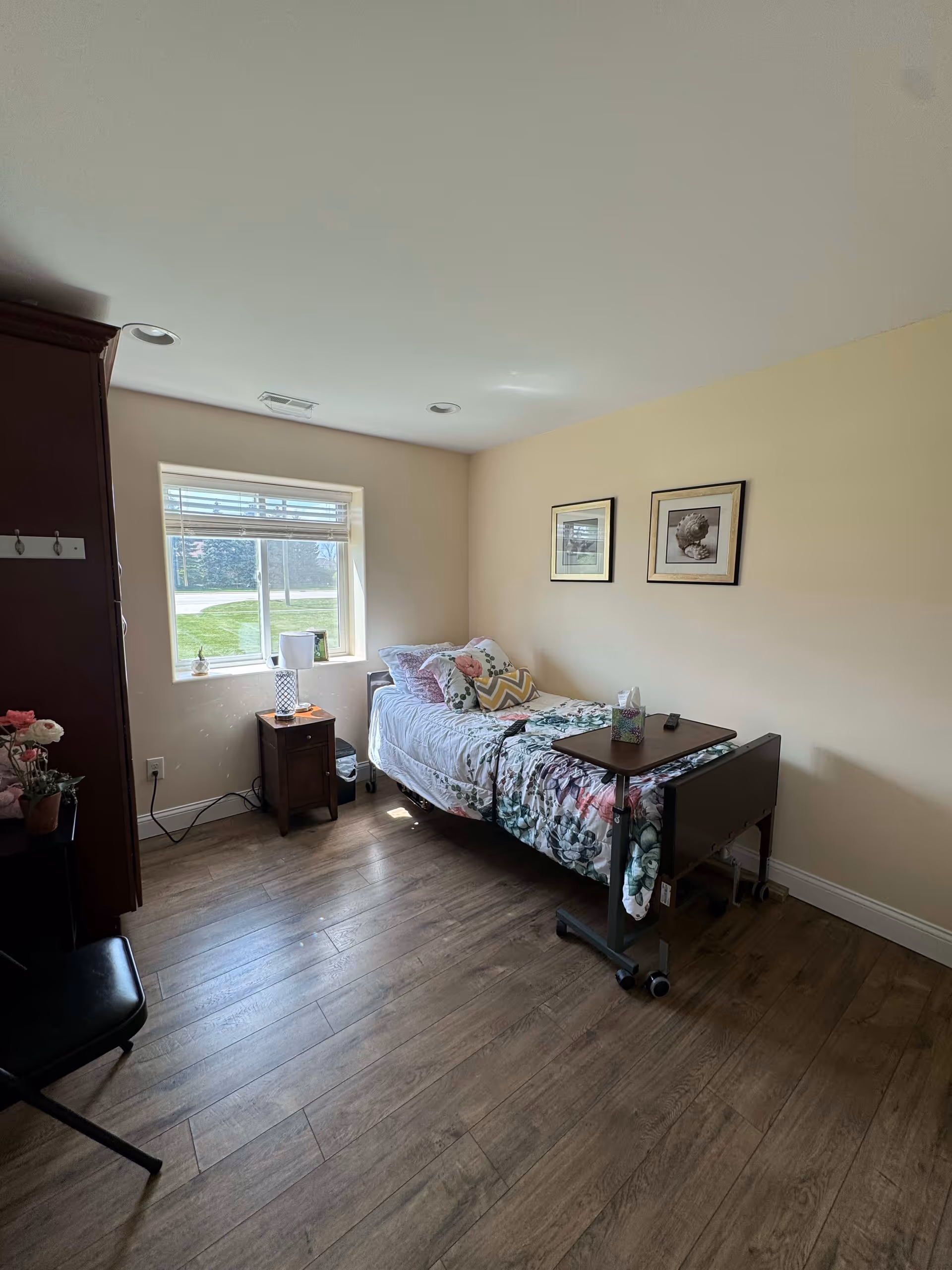 A small, cozy bedroom with a single bed covered in floral bedding and multiple pillows. There is a wooden bedside table with a lamp and a few items on it next to a window letting in natural light. The room has light-colored walls with two framed pictures hanging above the bed, wood flooring, and a dark wooden cabinet partially visible on the left side.
