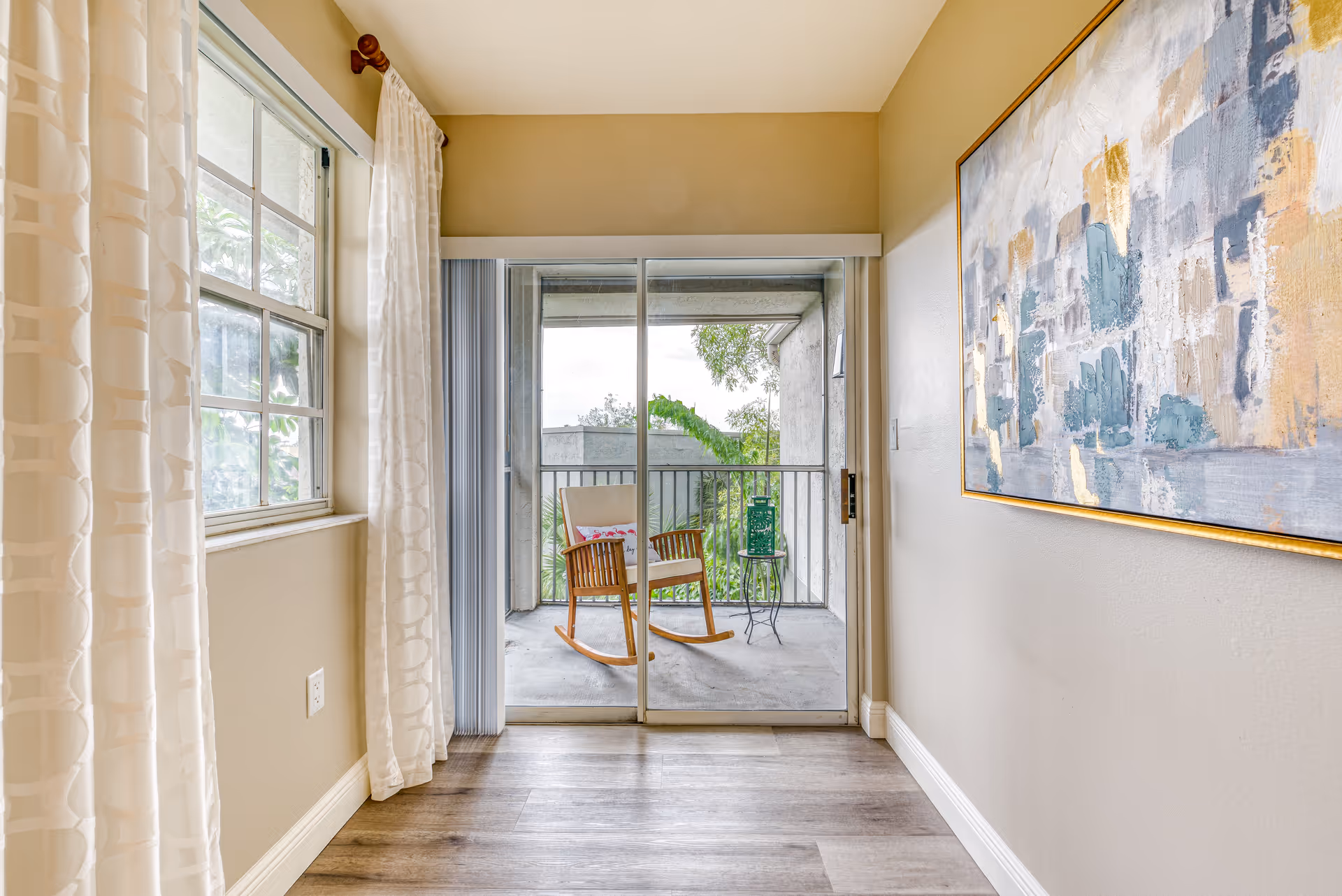 A bright interior hallway with wood flooring, beige walls, and a large abstract painting on the right wall. On the left, there is a window with white patterned curtains. At the end of the hallway, sliding glass doors open to a small balcony with a wooden rocking chair and a small round table with a green lantern. Trees and outdoor greenery are visible beyond the balcony.