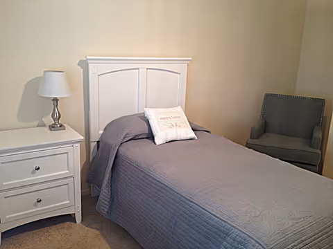 A small bedroom with a single bed covered in a gray quilt and a white pillow that says 'happy place'. Next to the bed is a white nightstand with two drawers and a silver lamp with a white lampshade. There is a gray upholstered armchair in the corner against a beige wall.