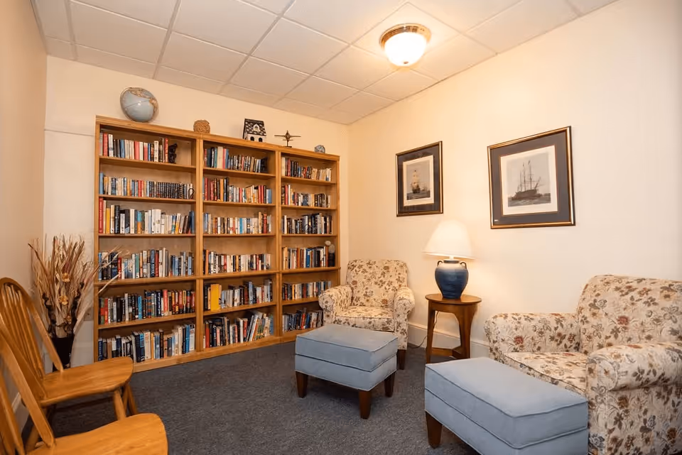 Cozy reading room with a large wooden bookshelf filled with books, two floral upholstered armchairs, two blue ottomans, a wooden side table with a blue lamp, two framed ship pictures on the wall, and two wooden chairs against the opposite wall.