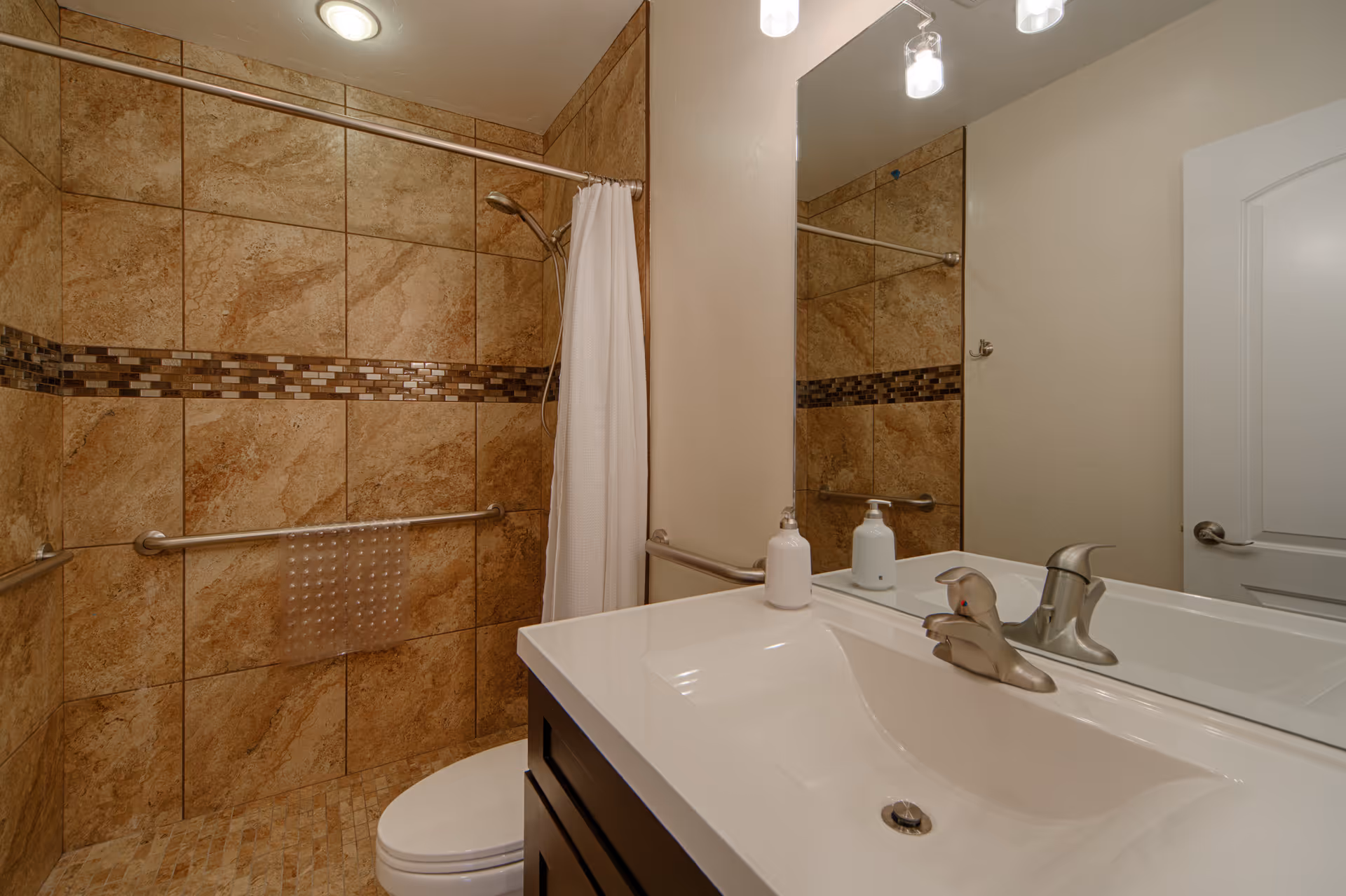 A bathroom with beige and brown tiled walls and floor, featuring a walk-in shower with a white curtain, a grab bar, and a textured plastic mat. There is a white sink with a silver faucet and two soap dispensers on the countertop. A large mirror is mounted above the sink, and a white door with a silver handle is visible in the background.