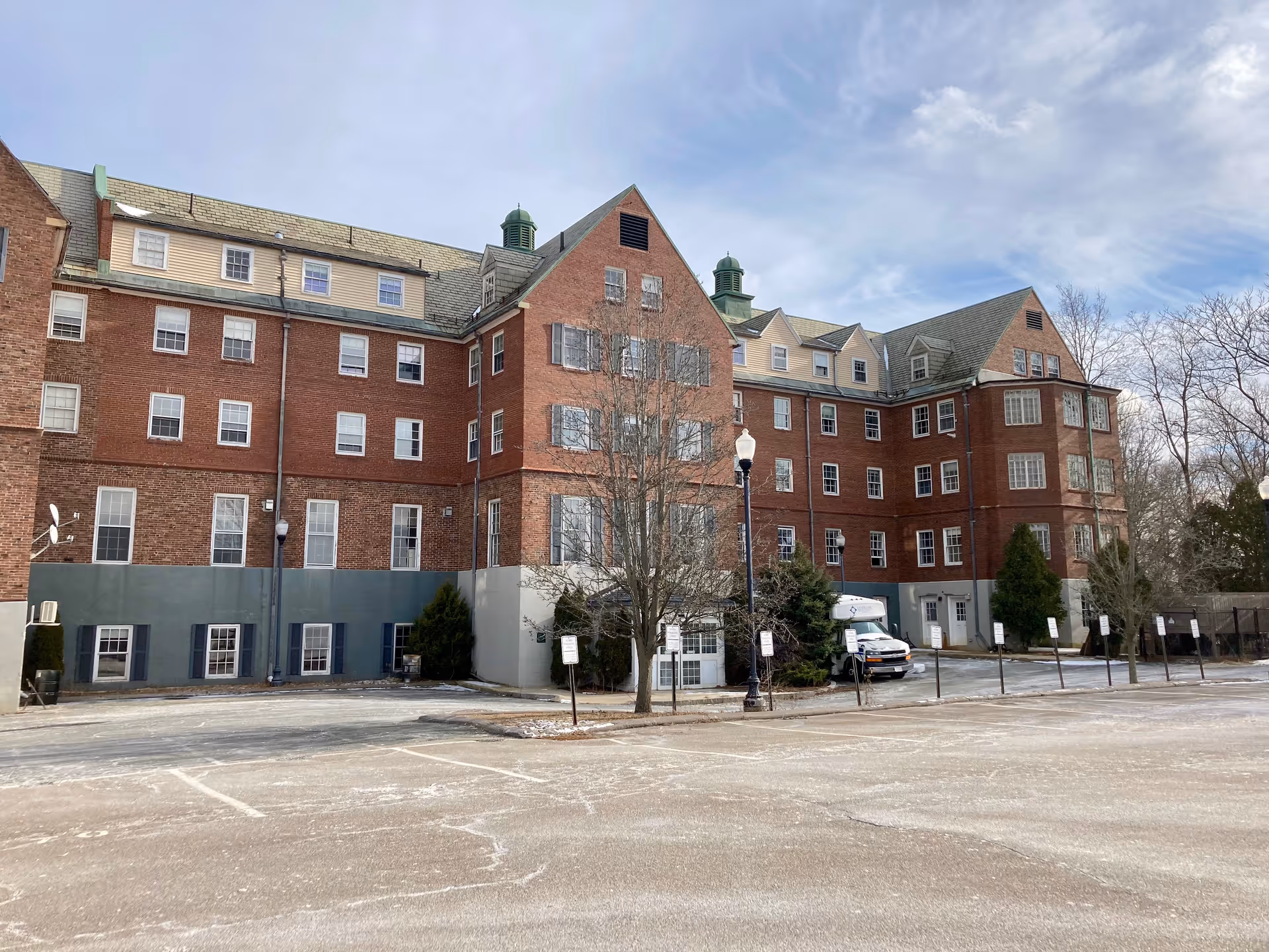 Exterior view of a large multi-story brick building with multiple windows and green cupolas on the roof. There is a parking lot in the foreground with several parking spaces and a few trees around the building. The sky is partly cloudy.