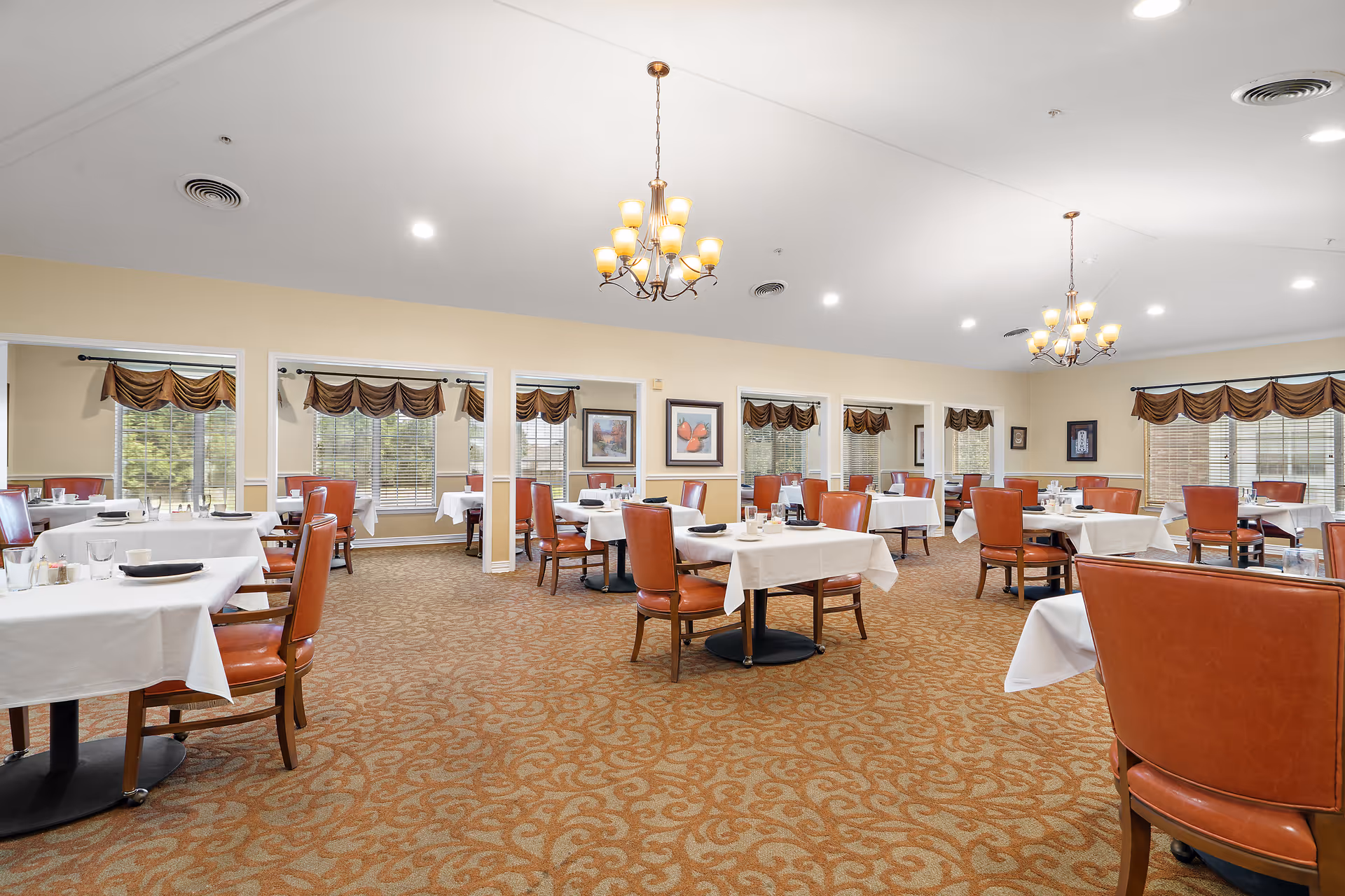 Spacious dining room with multiple tables covered in white tablecloths, each set with plates, glasses, and utensils. The room features brown patterned carpet, large windows with brown valances, and two chandeliers hanging from the ceiling. The walls are light yellow with framed artwork.