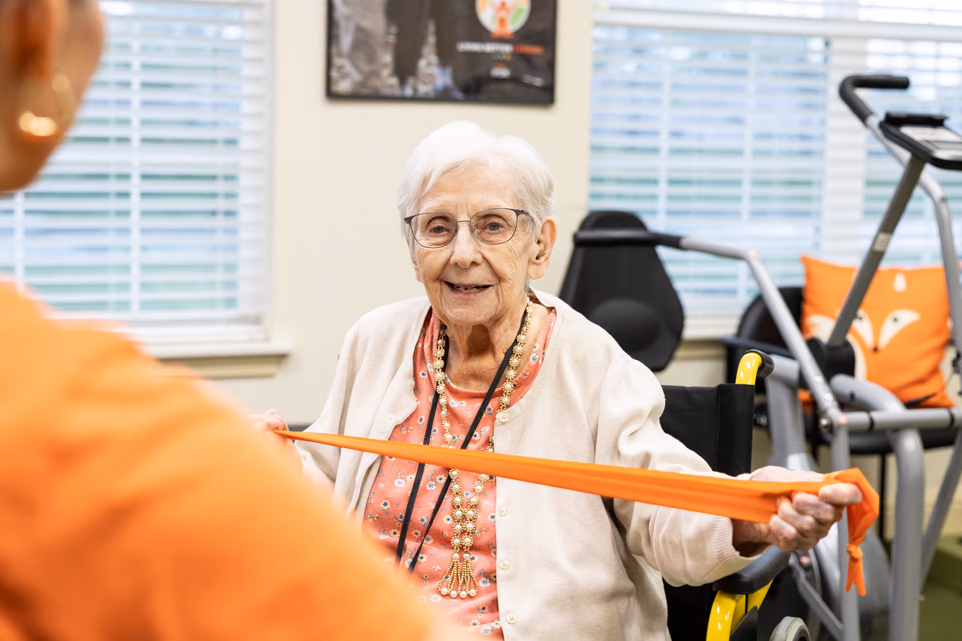 An elderly woman with white hair and glasses is seated in a wheelchair, smiling and holding an orange resistance band stretched between her hands. She is wearing a peach-colored floral top with a beige cardigan and a long beaded necklace. In the background, there is exercise equipment and a window with blinds.