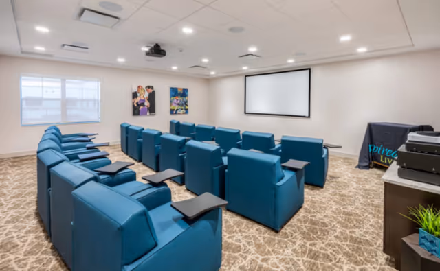 A small theater room with two rows of blue reclining chairs equipped with black tray tables, facing a large white projection screen on the wall. The room has beige patterned carpet, white walls, recessed ceiling lights, a projector mounted on the ceiling, and two colorful paintings on the wall near a window with blinds.
