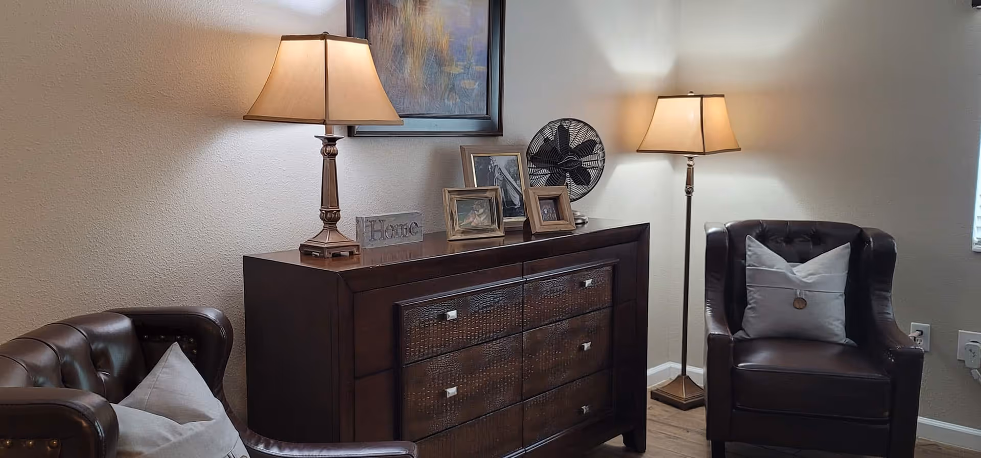 A cozy living room corner with two dark brown leather armchairs, each with a light-colored pillow. Between the chairs is a dark wooden dresser with six drawers, decorated with framed photos, a small decorative block with the word 'Home', and a table lamp. A floor lamp stands next to one of the armchairs, and a framed painting hangs on the wall above the dresser. The room has light-colored walls and wooden flooring.