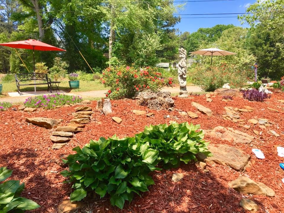 A landscaped garden area with red mulch, green plants, rocks, and bushes with red flowers. There are two patio umbrellas, one red and one beige, shading benches in the background. Trees and a clear blue sky are visible behind the garden.