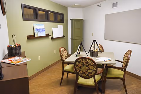 A small art room with a square table surrounded by four floral upholstered chairs. On the table are art supplies including paint palettes and brushes. The room has light green walls, a wooden floor, a small cabinet in the corner, and a shelf with canvases leaning against the wall.