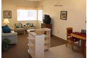 Small furnished living room with a sofa and two chairs, a TV on a stand, a bookshelf island and a small dining table near a window.