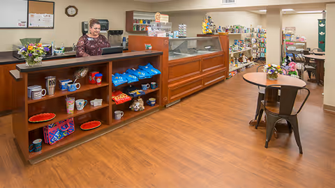 Interior view of a retail or reception area in a senior living facility. A woman stands behind a wooden counter with shelves displaying mugs, snacks, and small items. To the right, there is a glass display case and shelves stocked with various products. A round table with chairs and a flower arrangement is visible in the foreground. The floor is wooden, and the walls are beige with overhead lighting.