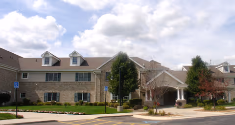 Exterior view of a senior living facility building with beige stone and siding, multiple windows, and dormer windows on the roof. The entrance is covered with a portico supported by white columns. There are landscaped bushes and trees around the building, a parking lot with handicap parking spaces, and a partly cloudy sky above.
