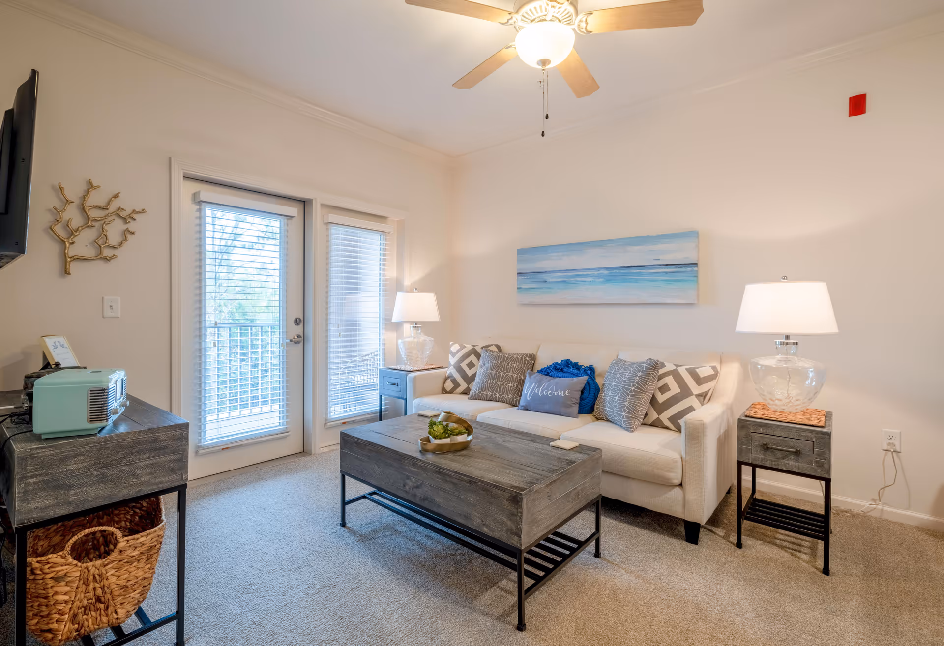 A cozy living room with a white sofa adorned with decorative pillows, a wooden coffee table with a small plant, two side tables each with a lamp, a ceiling fan with light, and a wall-mounted TV. There are glass doors with blinds leading to an outdoor balcony, and a piece of ocean-themed artwork hangs above the sofa.