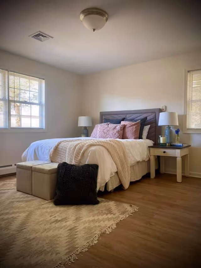 A cozy bedroom with a neatly made bed featuring white linens, a beige throw blanket, and multiple pillows in pink and dark blue. There are two bedside tables with matching lamps, a small blue vase with a flower, and a candle. The room has two windows with blinds, wooden flooring, a beige patterned rug, and two beige ottomans with a black knitted pillow in front of them.