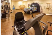 Indoor fitness room with exercise equipment including a recumbent bike and weight machines. A decorative vintage car is positioned against the wall, partially inside a painted tunnel mural. The room has wooden flooring and beige walls with bright overhead lighting.