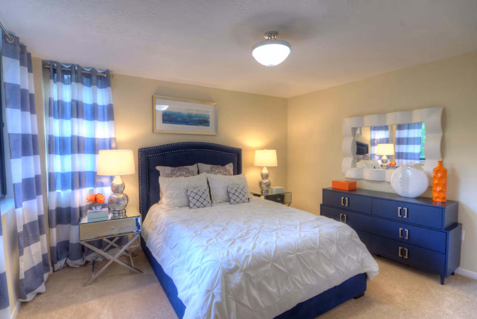 Well-lit bedroom with a navy-upholstered bed, patterned pillows, striped curtains, bedside lamps, and a navy dresser with decorative vases and a mirror.