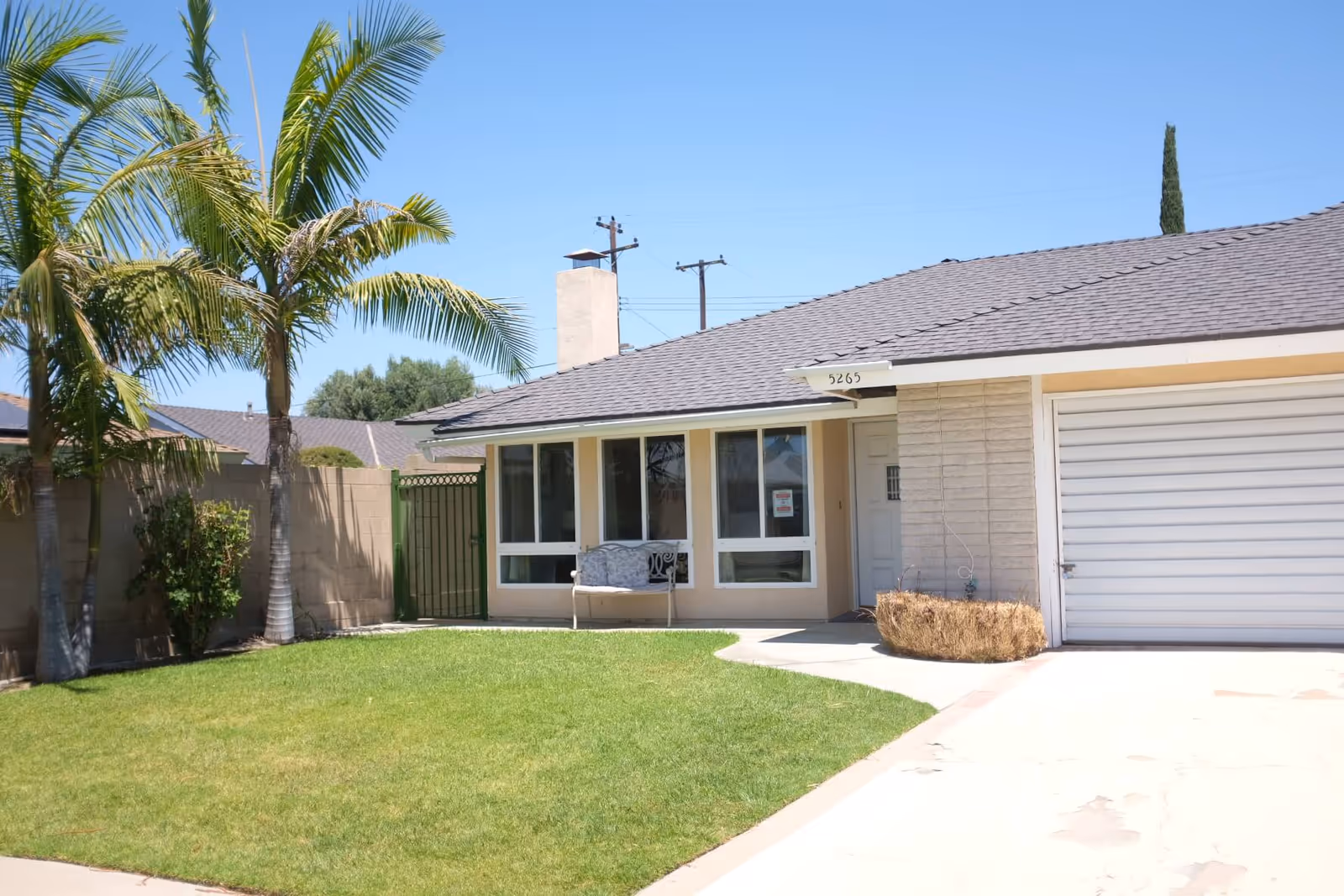 Front exterior view of a single-story house with a well-maintained green lawn, two palm trees, a bench near the window, and a closed white garage door under a clear blue sky.