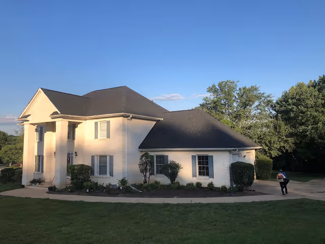 Exterior view of a two-story white brick building with a dark shingled roof, surrounded by green grass and trees under a clear blue sky. A person wearing a backpack is walking on the driveway to the right of the building.