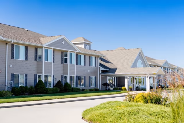 Exterior view of a two-story retirement community building with beige siding, multiple windows, a covered entrance, and well-maintained landscaping under a clear blue sky.