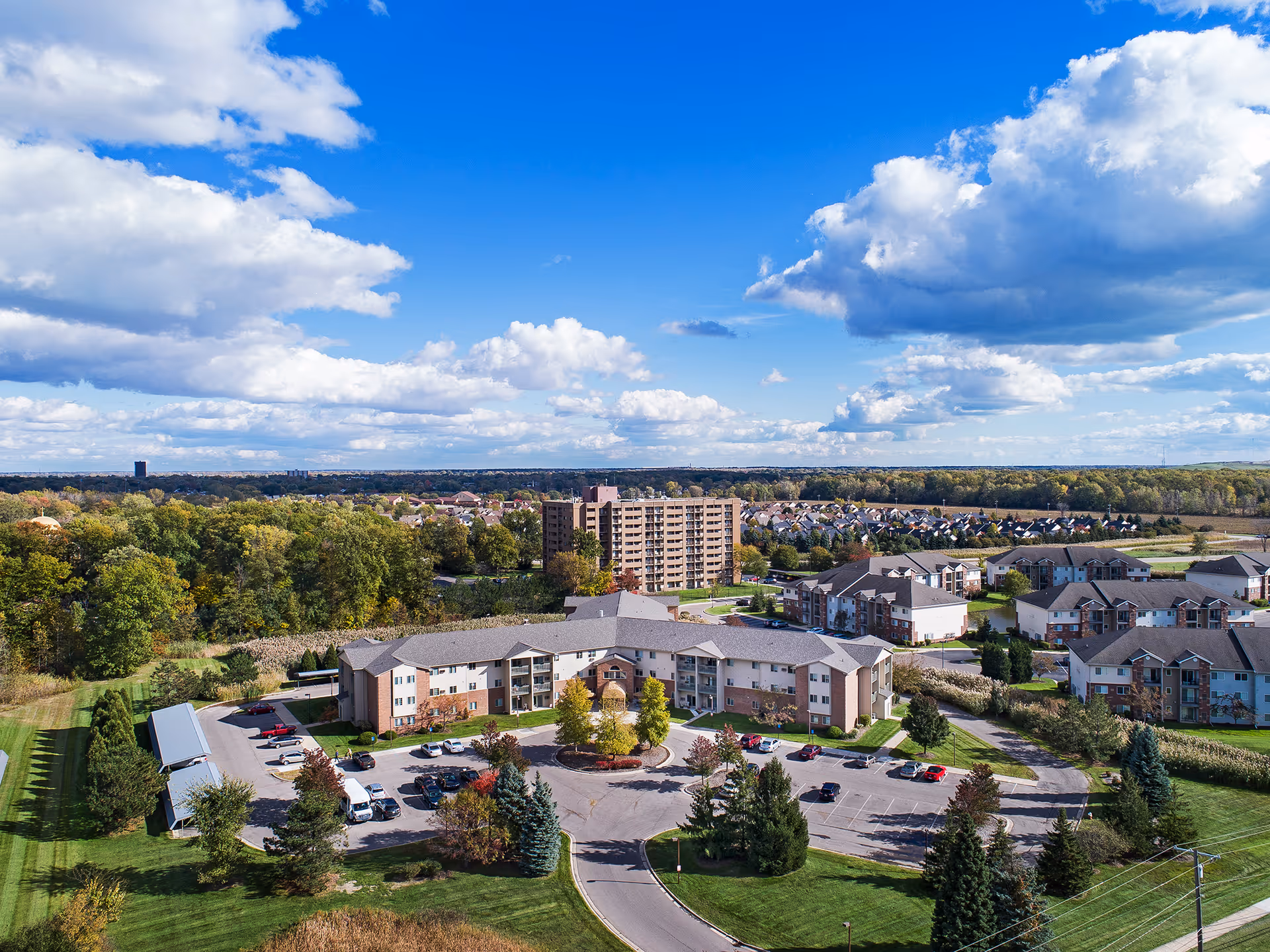 Aerial view of a senior living complex and surrounding residential neighborhood surrounded by trees under a blue sky.