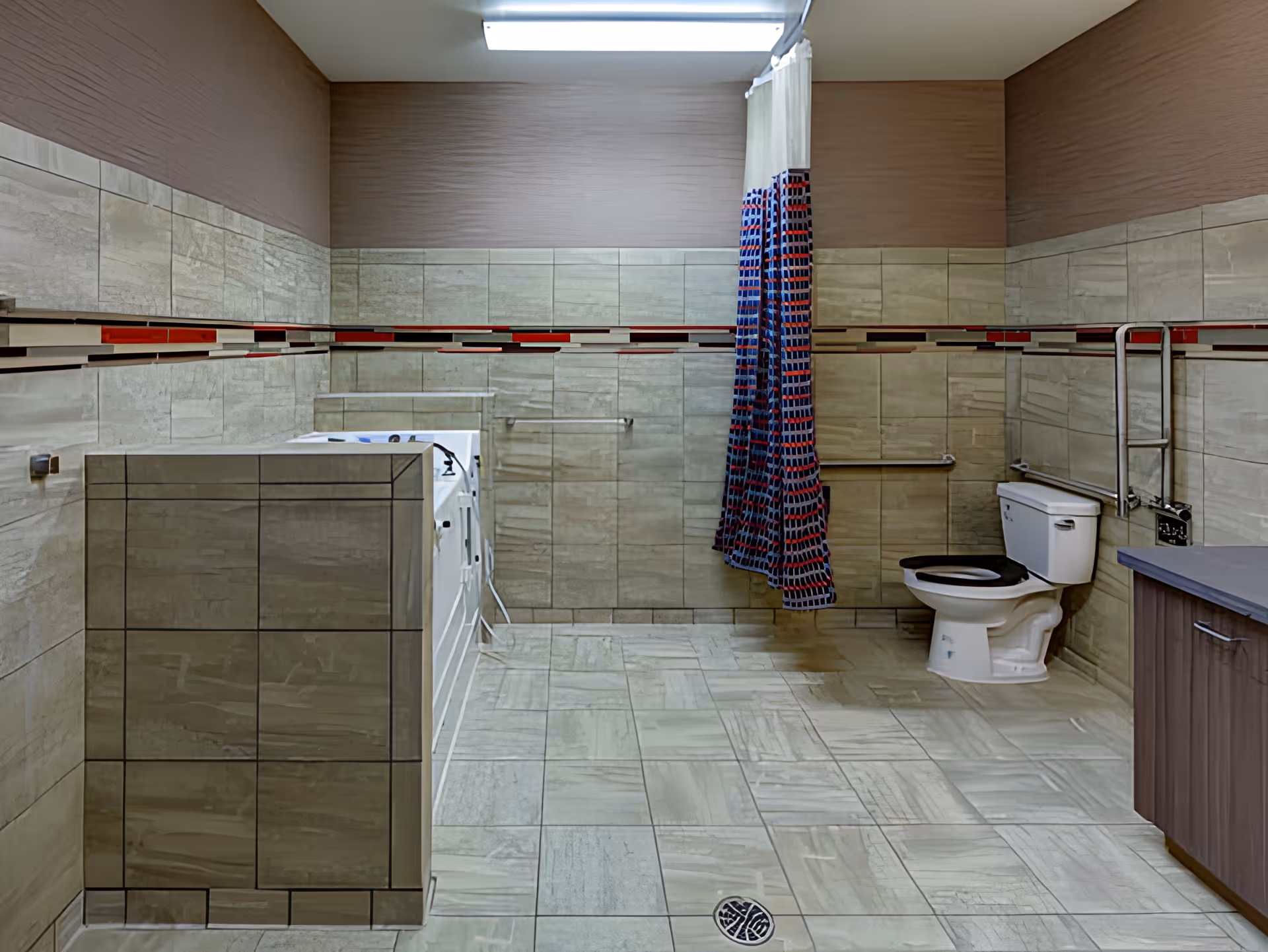 A spacious bathroom with beige tiled walls and floor, featuring a white toilet with a black seat, a bathtub with a colorful shower curtain, and a wooden cabinet with a gray countertop. The room has grab bars near the toilet and bathtub for accessibility.